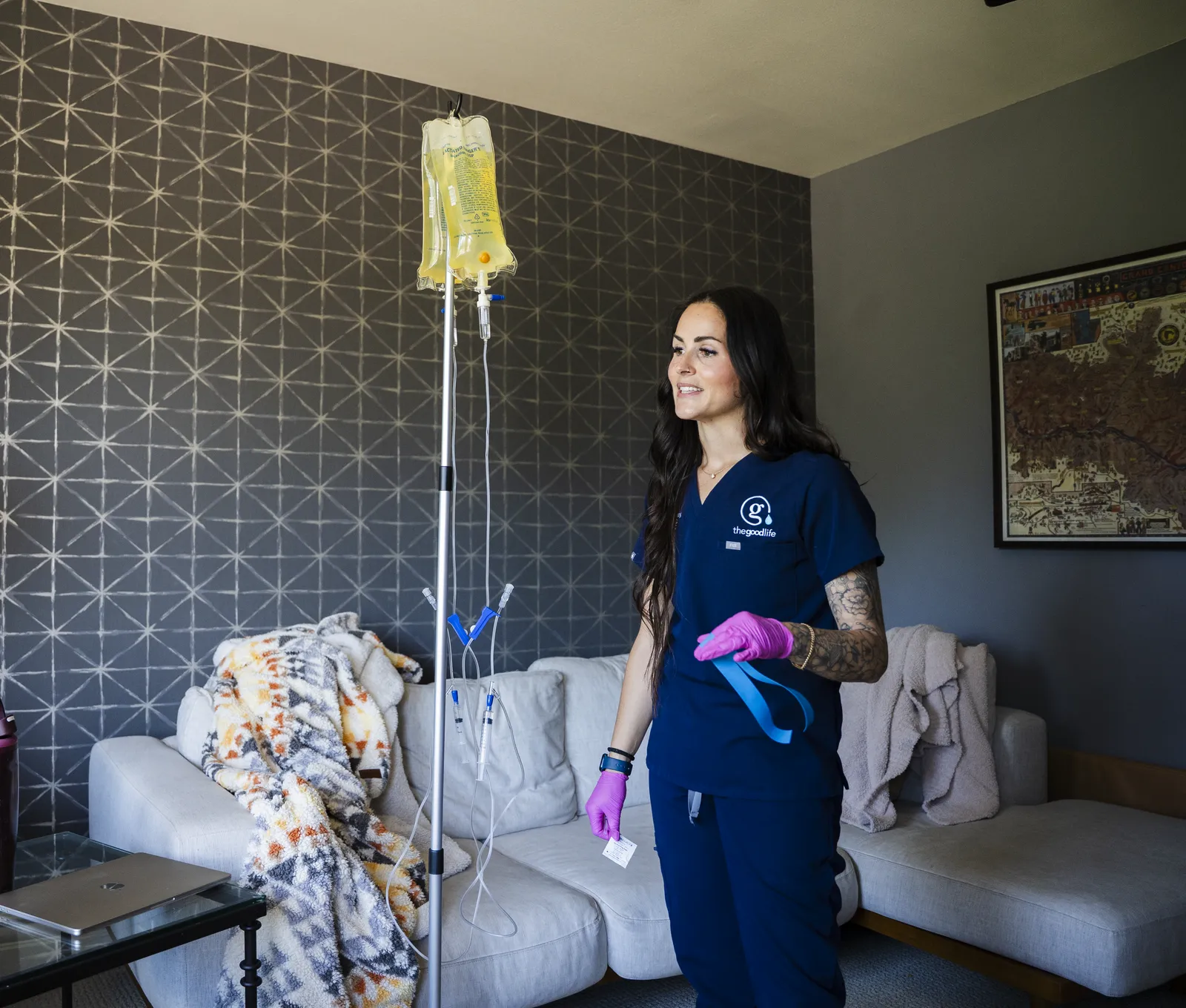 A Good Life nurse in navy scrubs smiling beside an IV drip in a client’s living room